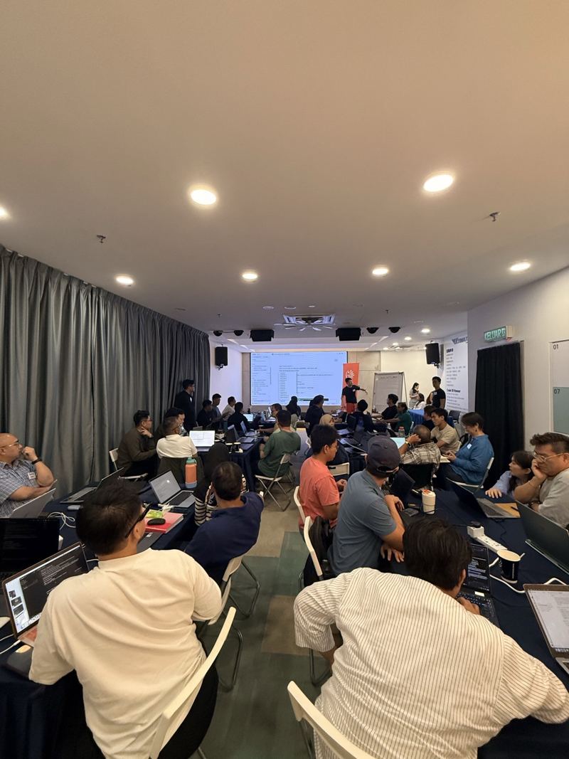 Attendees working on laptops at long tables while the presenter points at the projector screen